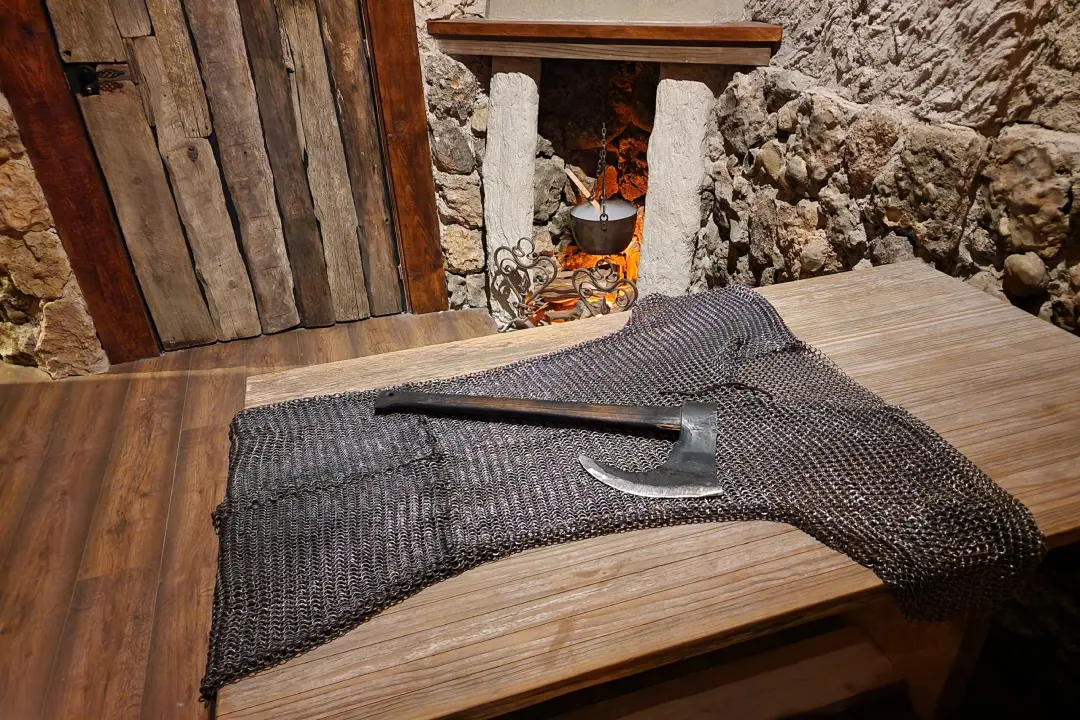 Armadura de malla de cota tendida sobre una mesa de madera con un hacha sobre ella y un caldero humeante de fondo